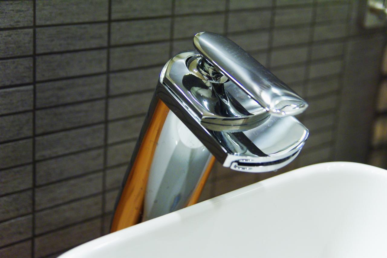Błyszcząca chromowana bateria umywalkowa z podniesionym uchwytem, widoczna na tle szarej, cegiełkowanej ściany i fragmentu białej umywalki.