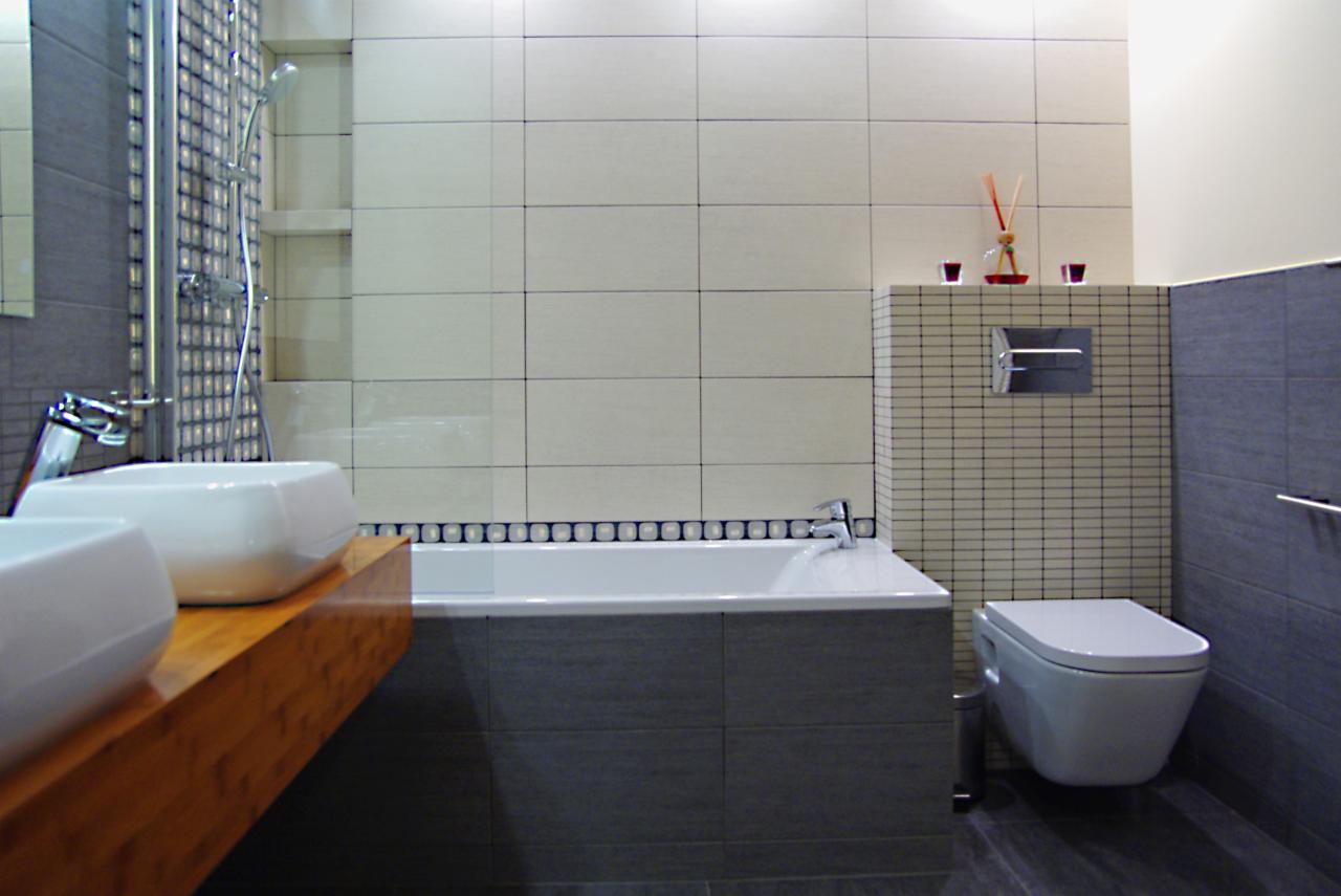 Nowoczesna łazienka z dwoma białymi umywalkami na drewnianym blacie, białą wanną obudowaną szarymi płytkami, toaletą wiszącą i prysznicem z deszczownicą. Na ścianach jasne płytki w dużym formacie...