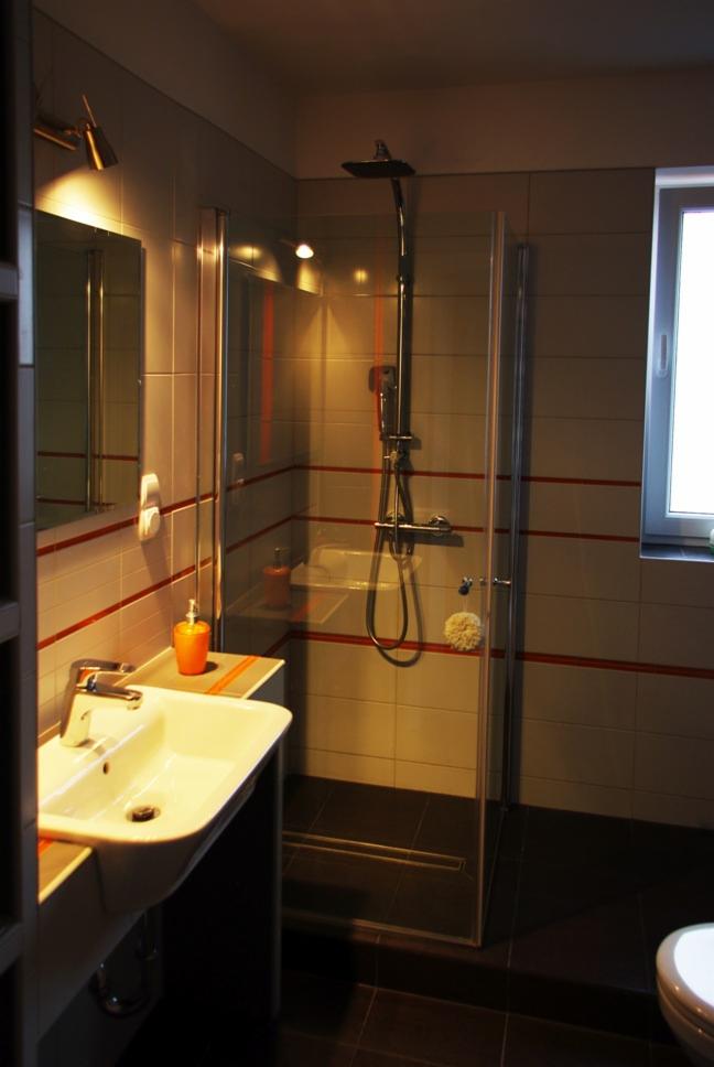 Wykończona łazienka w Poznaniu: biała umywalka z baterią, szklana kabina prysznicowa z deszczownicą, beżowe płytki z czerwonym paskiem, czarna podłoga i okno z białymi ramami.