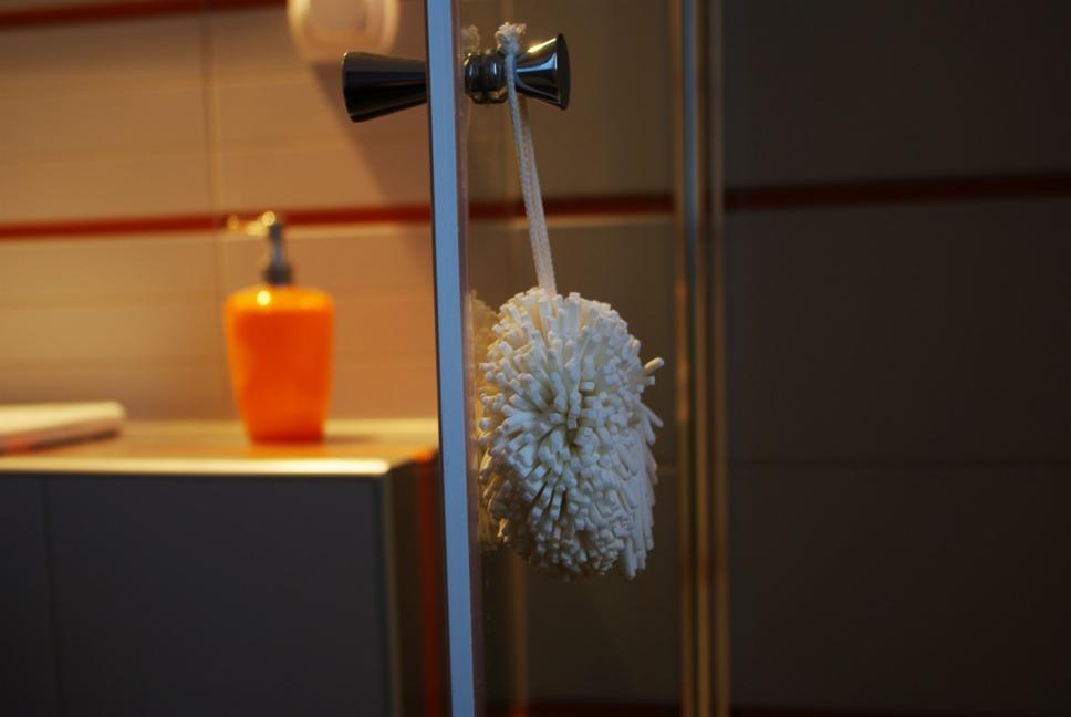 Wnętrze łazienki w Poznaniu: biała myjka zawieszona na szklanej kabinie prysznicowej, w tle pomarańczowy dozownik do mydła i płytki w ciepłych odcieniach.