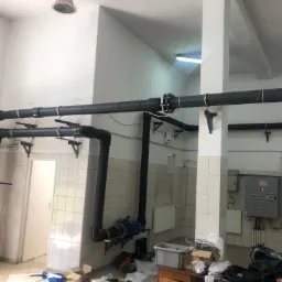 Czarne rury instalacyjne z zaworami i elementami łączeniowymi, zamocowane na ścianie wyłożonej kafelkami, widoczne szafy sterownicze, bałagan na stole.