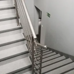 Schody pokryte szarą wykładziną z ciemniejszymi pasami, z metalową balustradą i znakiem ewakuacyjnym na szarej ścianie.