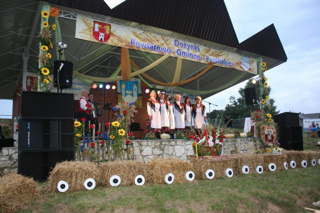 Scena dożynkowa z nagłośnieniem, dekoracjami z kwiatów i siana. Na scenie zespół ludowy w strojach regionalnych i muzyk z akordeonem.