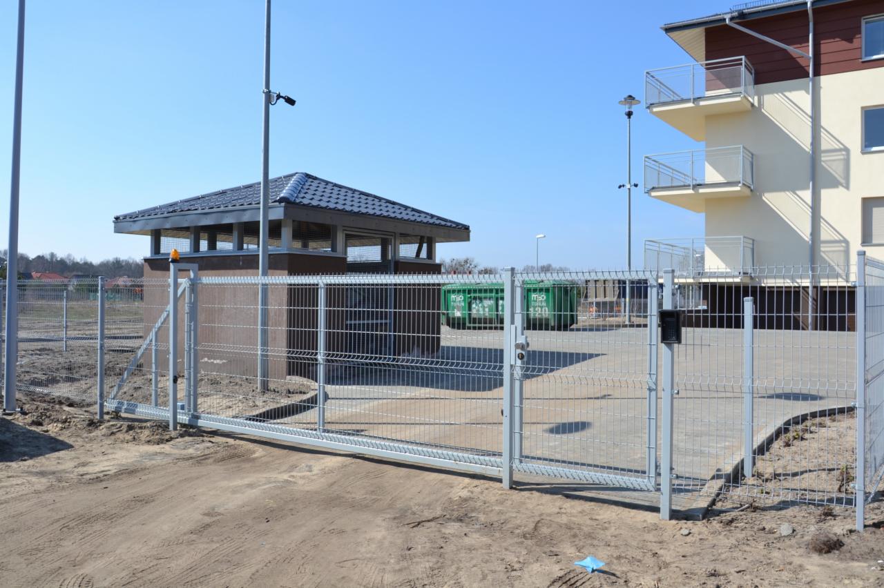 Przesuwna brama z siatki panelowej na wjeździe, obok budka strażnicza, budynek z balkonami w tle. Widoczne kosze na śmieci i oświetlenie zewnętrzne.