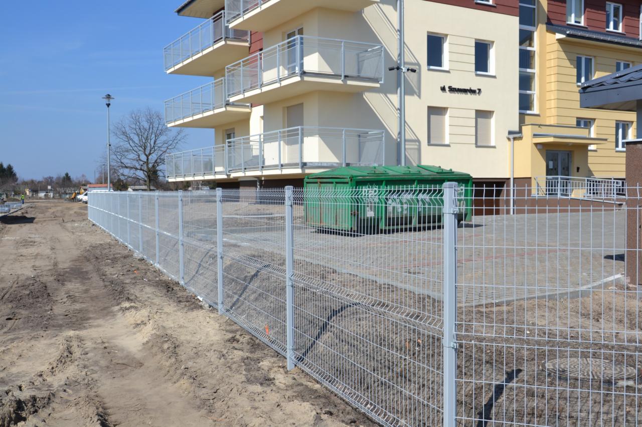 Nowoczesne, szare ogrodzenie panelowe z widocznym budynkiem mieszkalnym w tle i zielonym kontenerem na śmieci. Kątowe ujęcie z perspektywy ziemi.