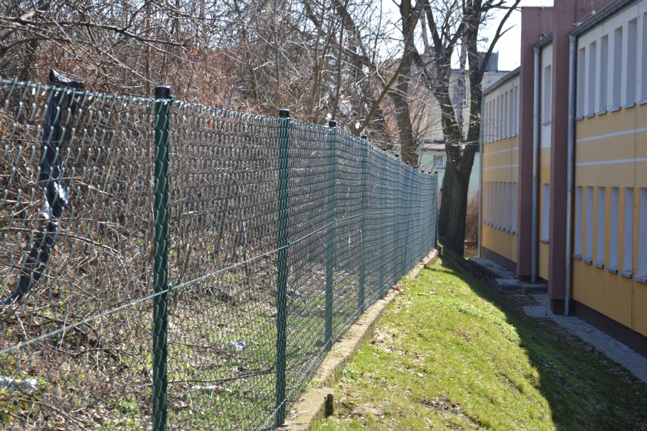 Zielone ogrodzenie z siatki, z widoczną podmurówką, oddzielające trawnik od zarośli i budynku z żółtą elewacją w tle.