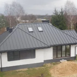 Dom jednorodzinny z szarym dachem blaszanym z dwoma oknami dachowymi i ciemnymi rynnami, na tle drzew i pochmurnego nieba. Widoczny fragment elewacji i sterta piasku.