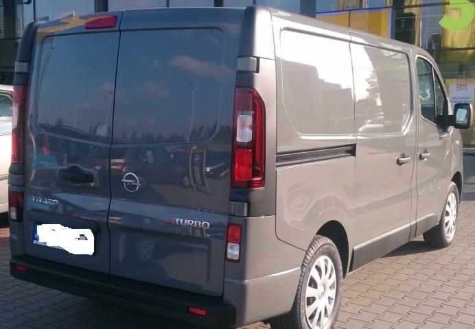 Szary bus Opel Vivaro z widocznym napisem 'Turbo' na tylnej klapie, zaparkowany na zewnątrz budynku z odblaskową szybą.