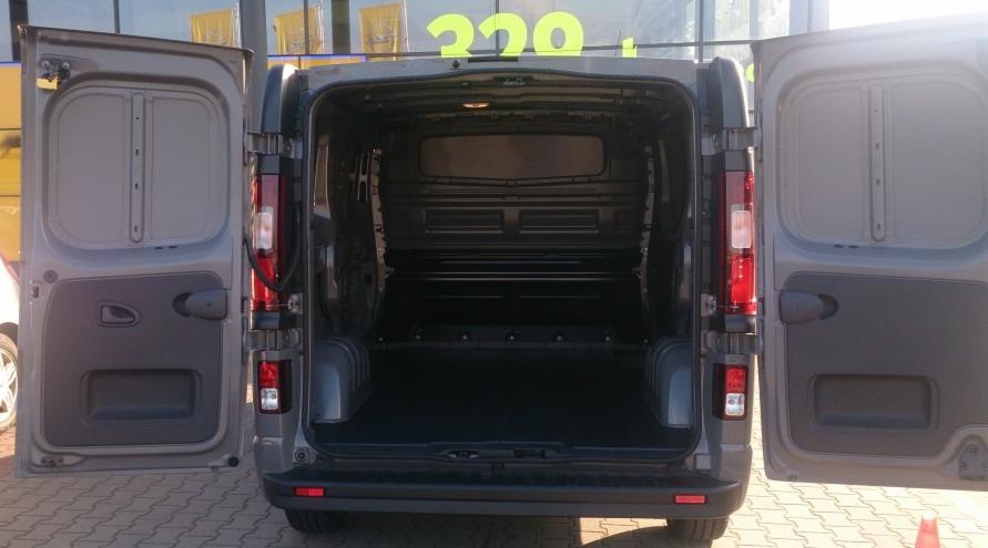 Otwarty, pusty bagażnik szarego busa marki Opel Vivaro, widok z tyłu na kostkę brukową i budynek z żółtym szyldem.