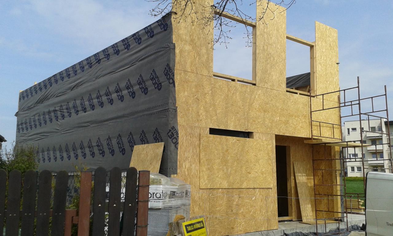 Wznoszenie konstrukcji domu z drewnianych paneli OSB, widoczne elementy szkieletu budynku, okna bez szyb, czarna membrana wiatroizolacyjna z nadrukiem, rusztowanie przy ścianie bocznej oraz paleta...