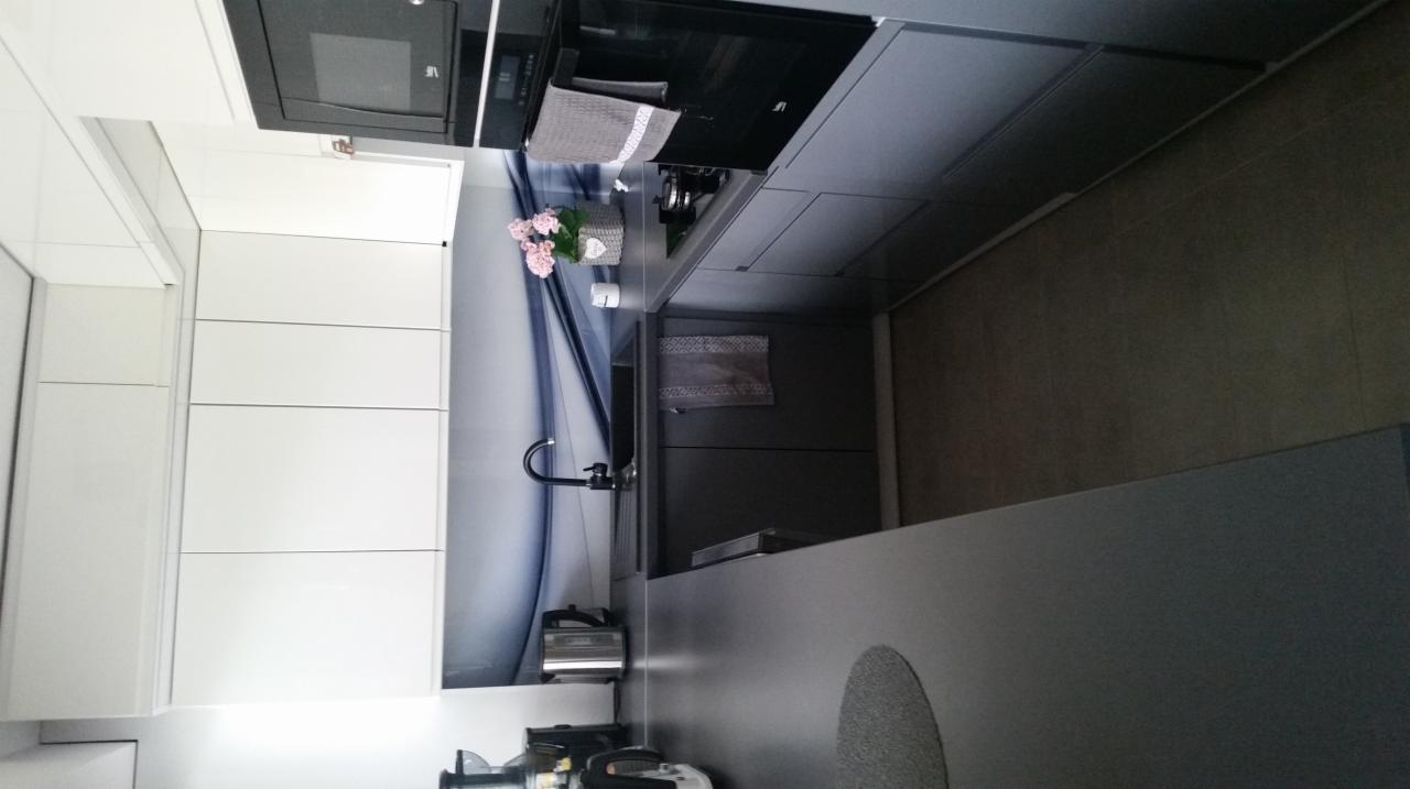 Nowoczesna kuchnia z białymi szafkami, szarym blatem i panelem ściennym z motywem wodnym, uzupełniona o kwiaty w ozdobnym koszyku i kuchenne akcesoria.