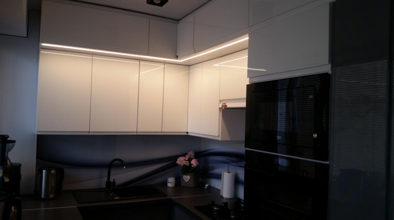 Nowoczesna, minimalistyczna kuchnia z białymi, błyszczącymi szafkami podświetlonymi listwami LED, czarnym zlewem i baterią oraz piekarnikiem w zabudowie.