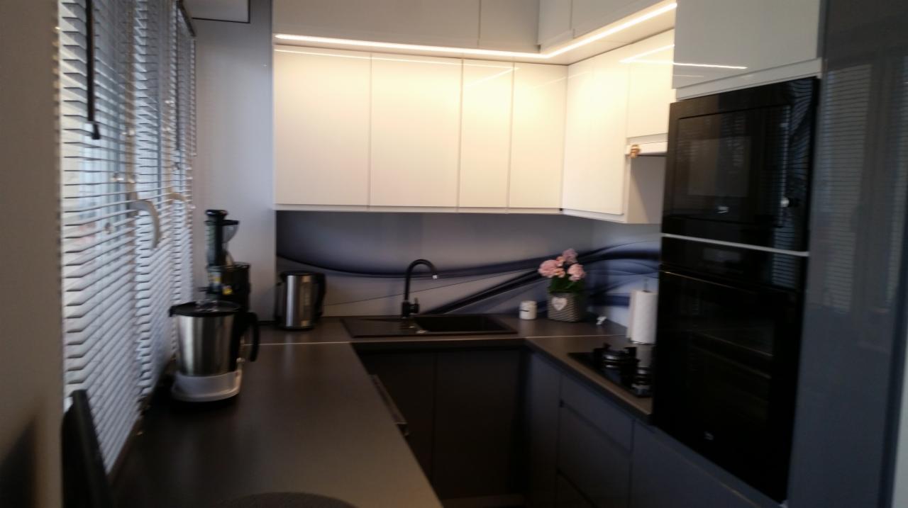 Nowoczesna kuchnia z białymi szafkami górnymi podświetlonymi LED i szarymi szafkami dolnymi, czarnym zlewem i baterią na tle grafiki ściennej, zabudowana piekarnikiem i płytą grzewczą, widoczne...