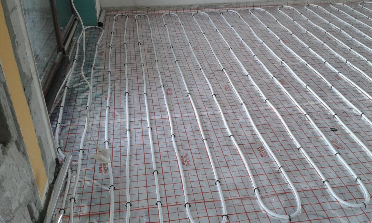 Białe rurki ogrzewania podłogowego ułożone na srebrnej folii z czerwoną siatką, widoczne czarne uchwyty mocujące rurki.