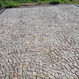 Ułożona kostka granitowa w łukowaty wzór, widok z góry, z zielenią w tle.