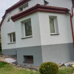 Dwukolorowa elewacja domu z czerwonym dachem i rynnami. Jasnoszara g&oacute;ra kontrastuje z ciemniejszym dołem. Widoczne okna i fragment ogrodu z krzewem.