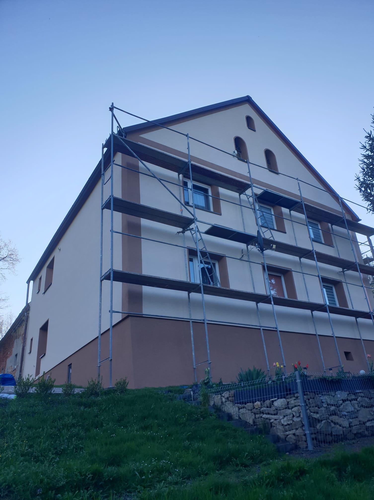 Dwukolorowa elewacja domu w trakcie prac, z rusztowaniem. Jasnobeżowa góra i brązowy dół, zieleń trawy i błękit nieba w tle. Ujęcie z dołu.