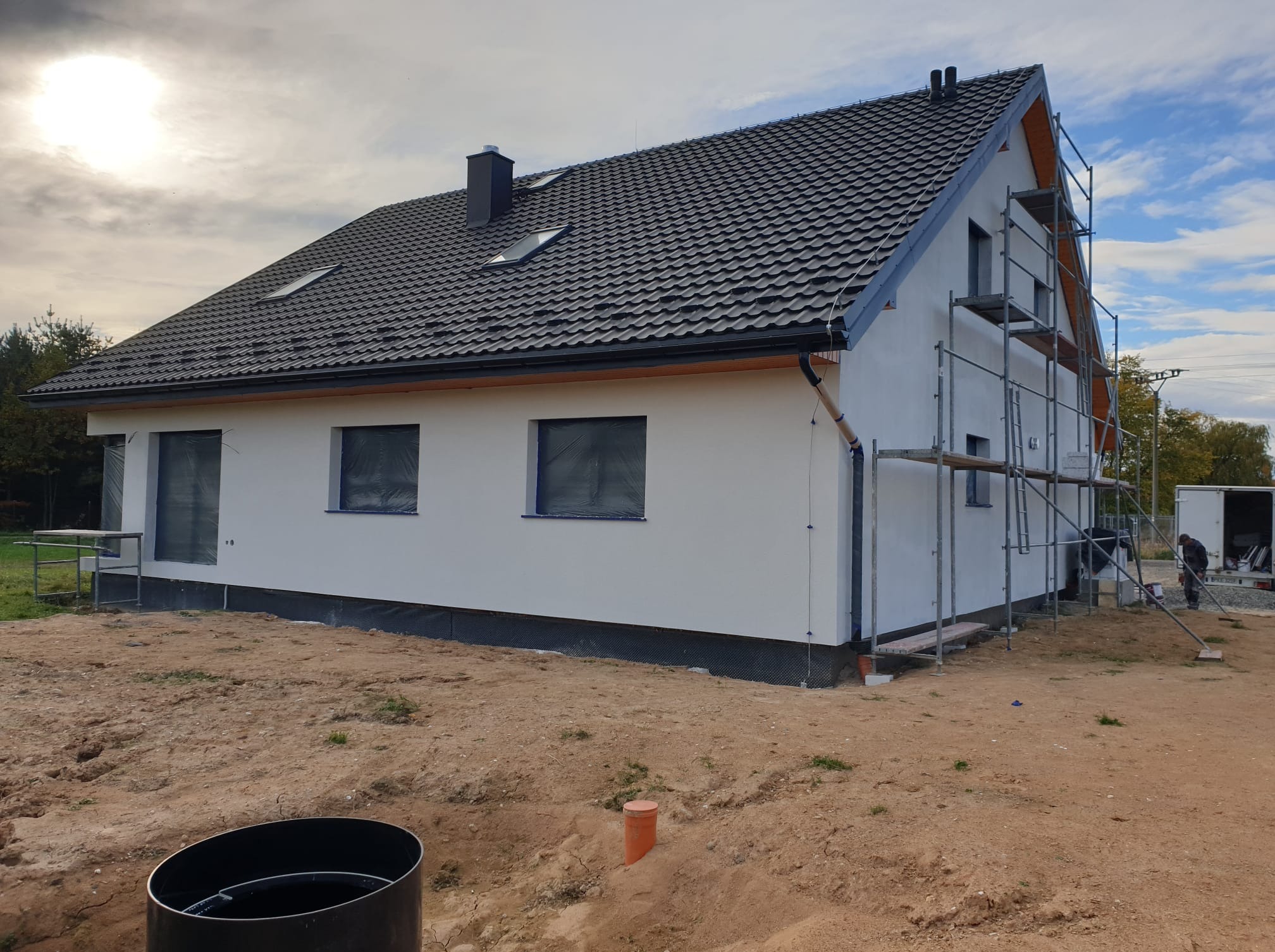 Biały dom jednorodzinny z ciemną dachówką, okna zabezpieczone folią, rusztowania przy ścianie bocznej, teren wokół w trakcie prac wykończeniowych.