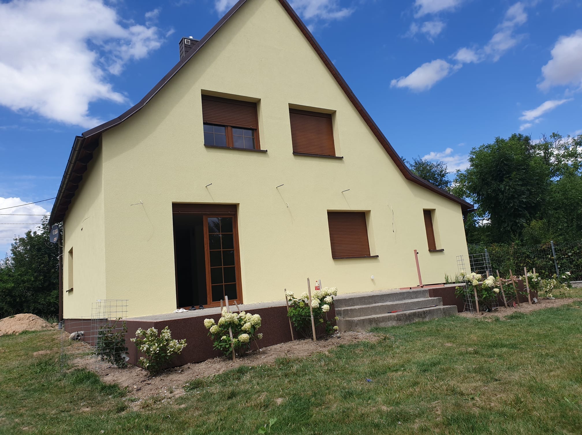 Jasnożółty dom jednorodzinny z brązowymi oknami i roletami, widok z ogrodu. Świeżo pomalowana elewacja, widoczne schody wejściowe i rabaty kwiatowe.