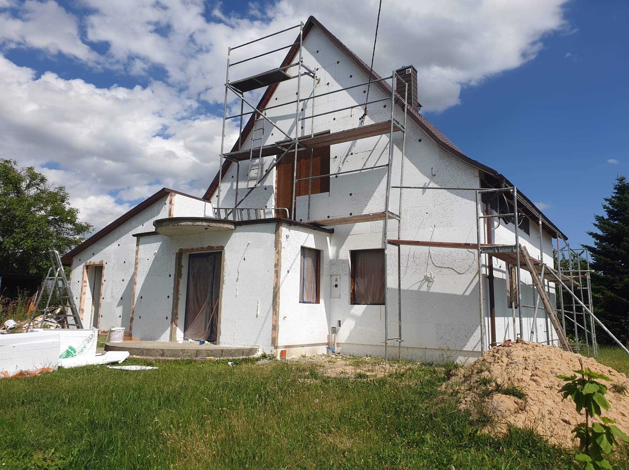 Dom jednorodzinny w trakcie ocieplania styropianem, z widocznymi rusztowaniami i przygotowanymi materiałami izolacyjnymi. Słoneczny dzień, częściowo zachmurzone niebo.