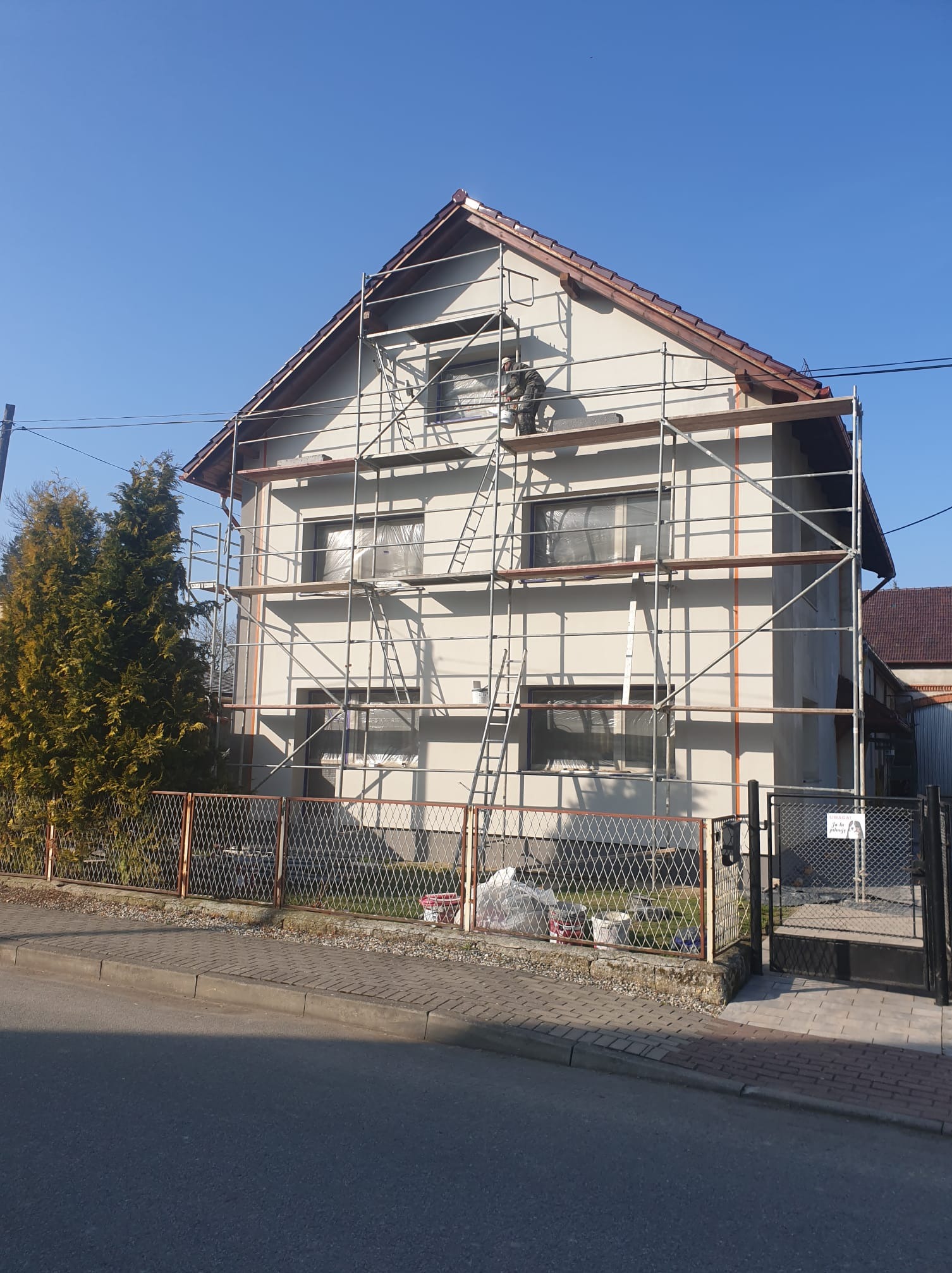 Malowanie elewacji domu jednorodzinnego z rusztowania. Okna zabezpieczone folią, widoczne puszki z farbą. Pracownik w kombinezonie maluje ścianę. Słoneczny dzień.