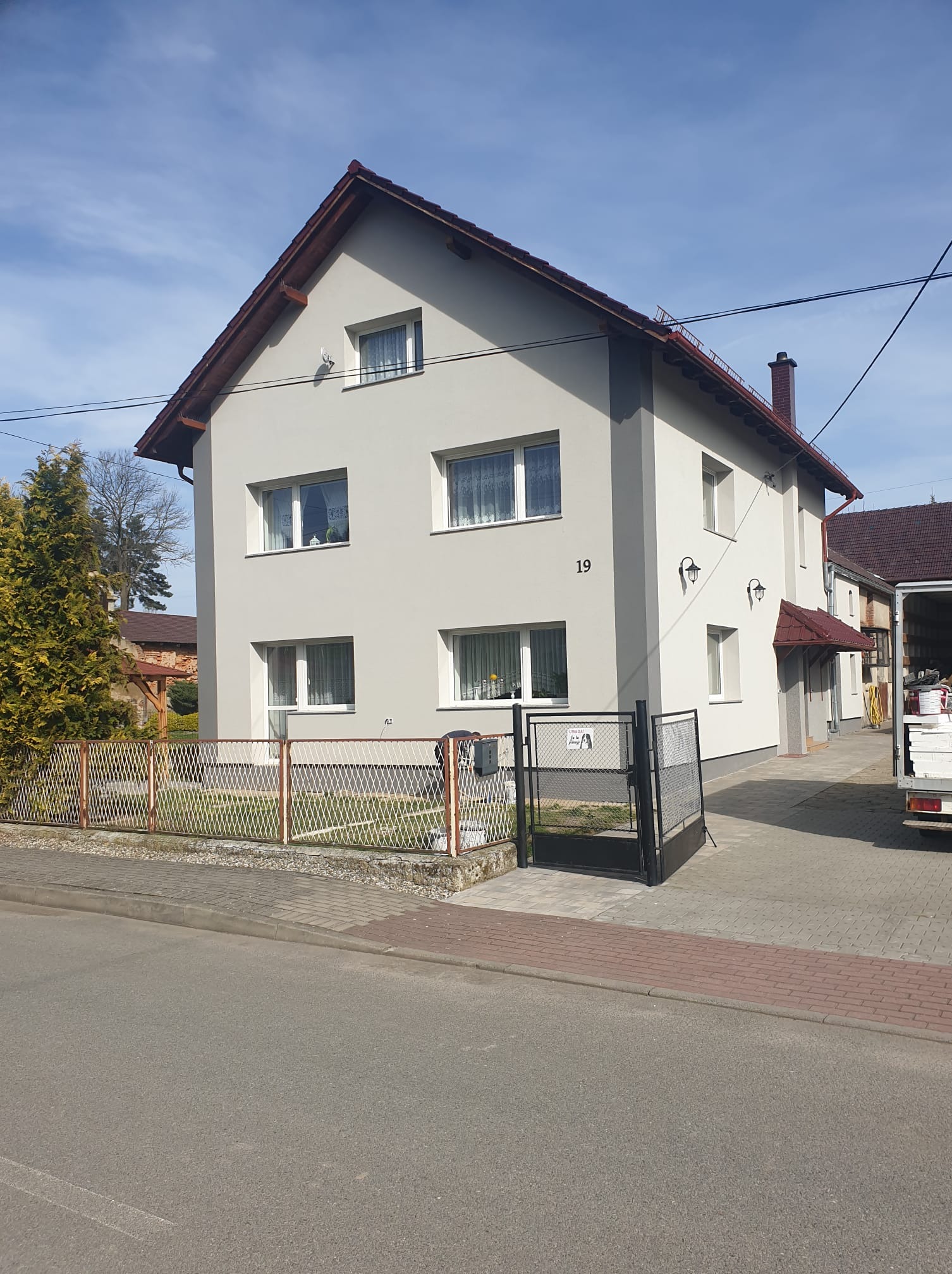 Dwupiętrowy dom z jasną elewacją i ciemnym dachem, numer 19 na ścianie, ogrodzony metalową siatką. Widoczne lampy zewnętrzne i fragment samochodu dostawczego.