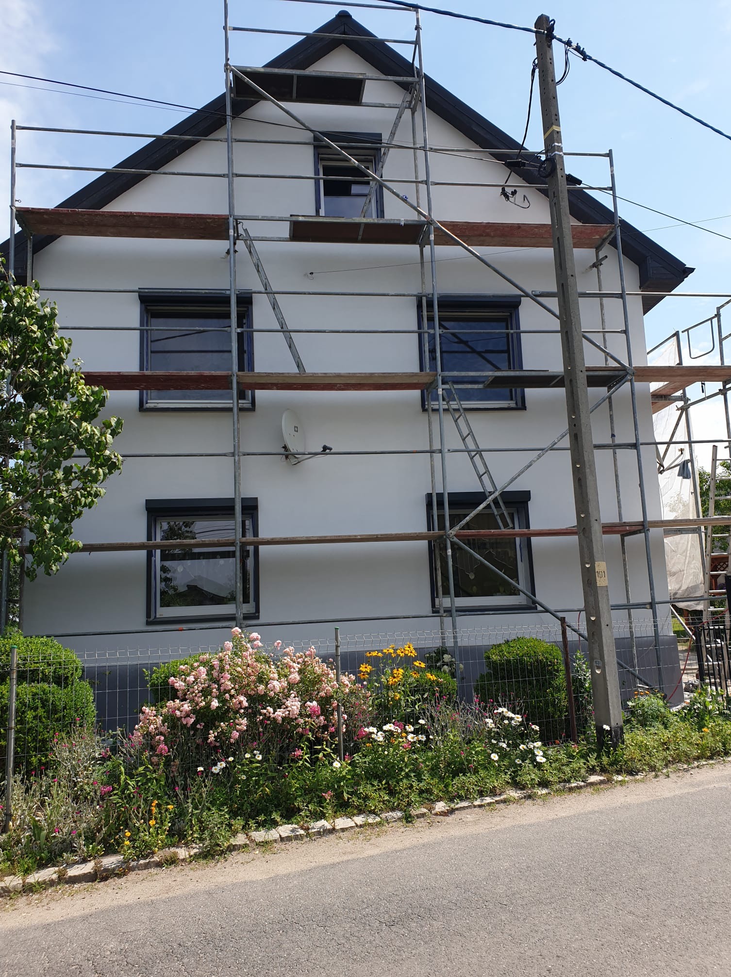 Dom jednorodzinny w rusztowaniach, świeżo pomalowana elewacja w jasnym odcieniu, okna z ciemnymi ramami. W tle błękitne niebo i zieleń drzew.