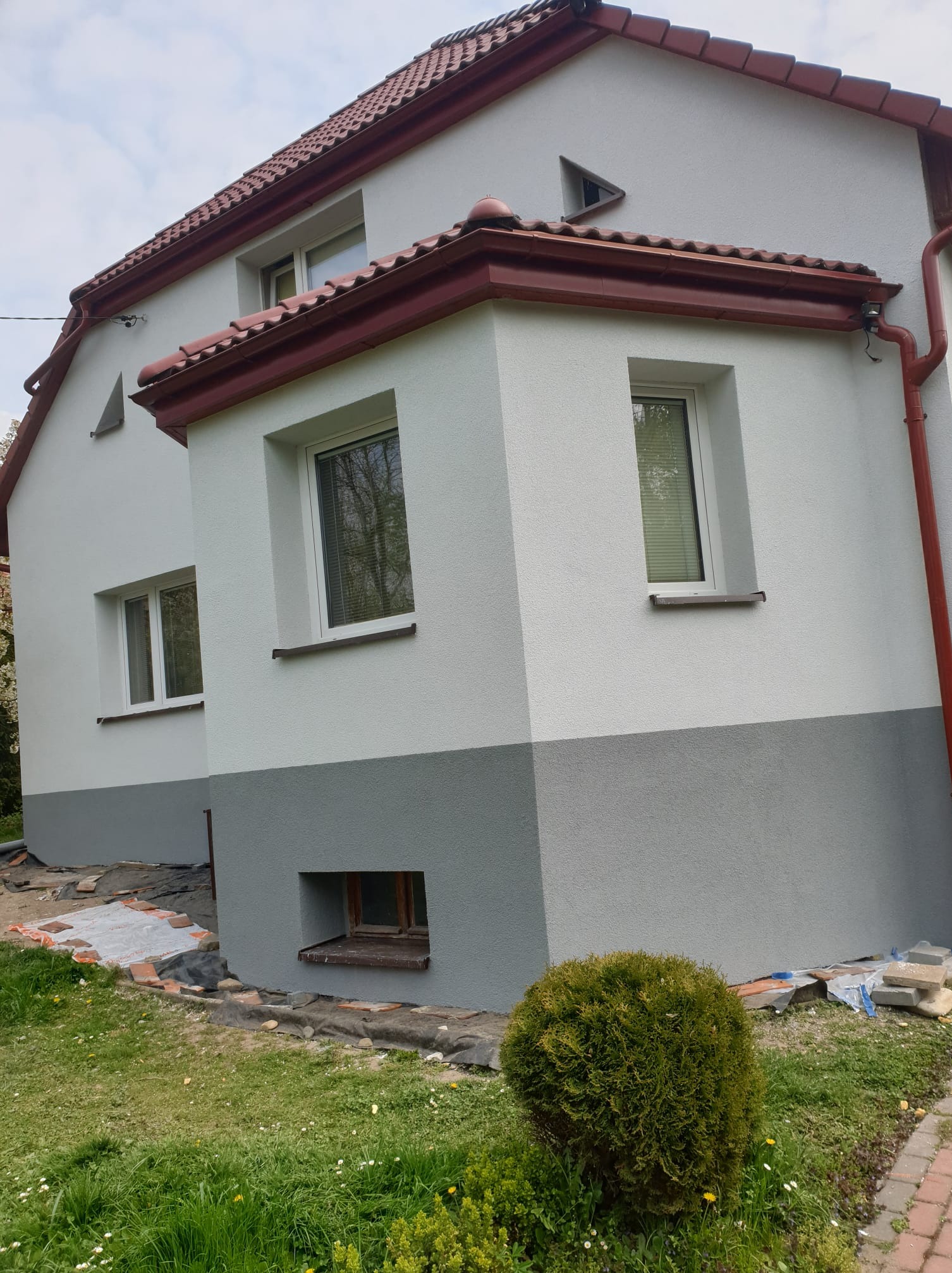 Dwukolorowa elewacja domu z czerwonym dachem i rynnami. Jasnoszara góra kontrastuje z ciemniejszym dołem. Widoczne okna i fragment ogrodu z krzewem.