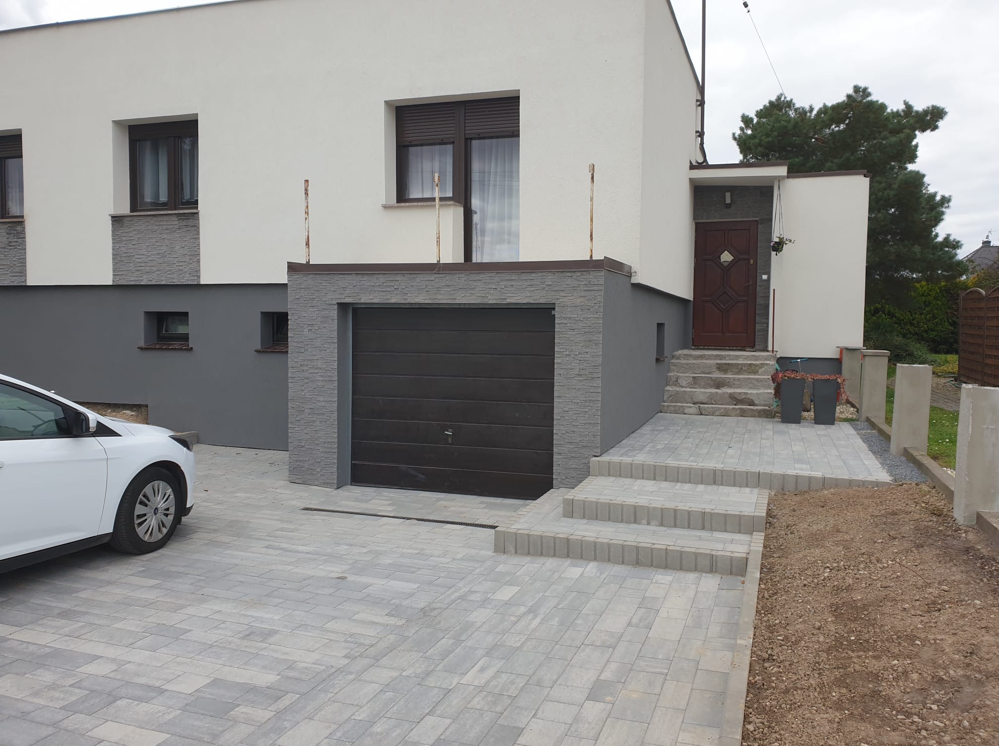 Dwukondygnacyjny dom z garażem i elewacją w odcieniach bieli i szarości. Widoczne schody wejściowe i podjazd z kostki brukowej. Częściowo widoczny biały samochód.
