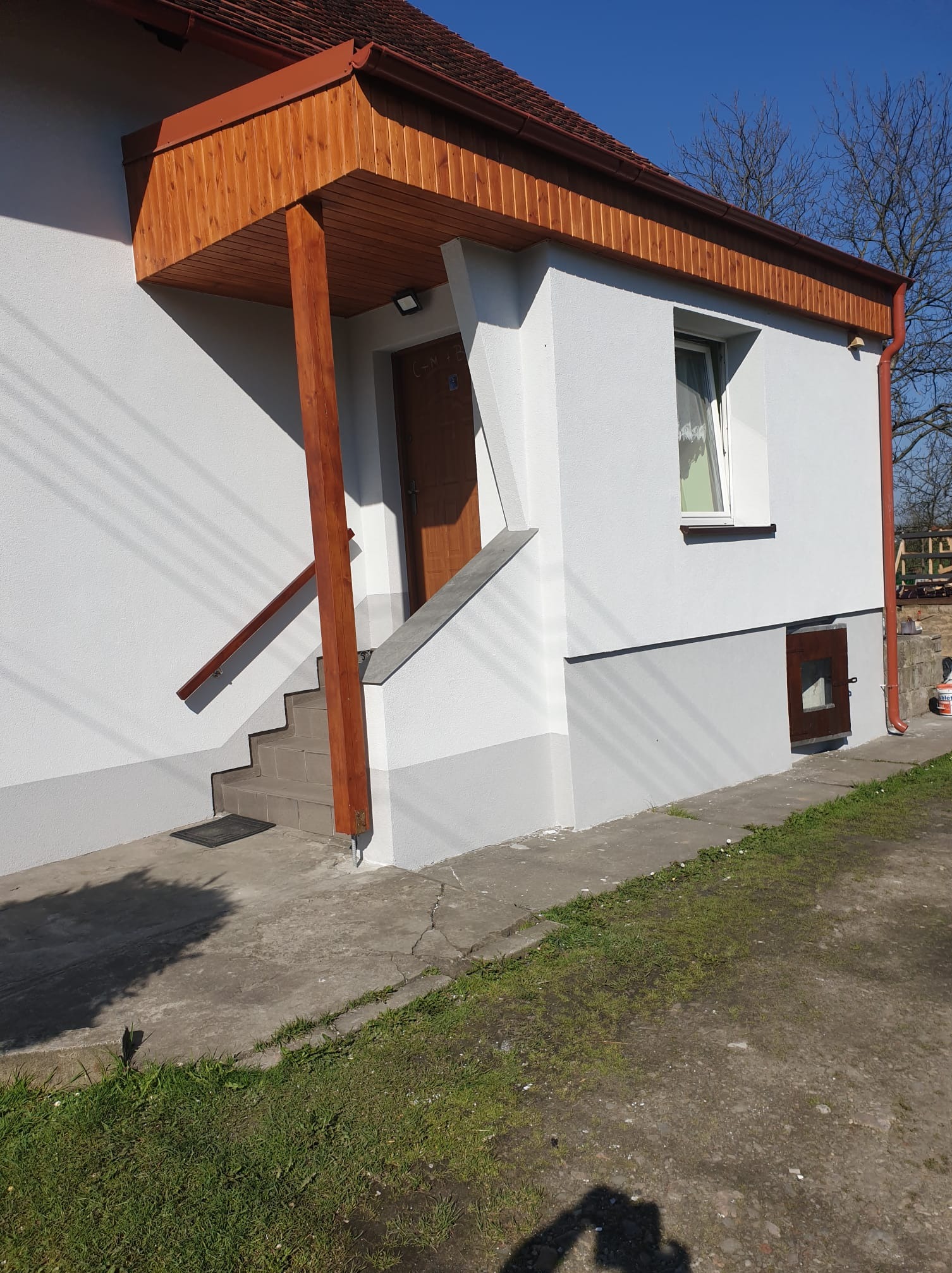 Biała elewacja domu z drewnianym zadaszeniem nad wejściem i szarą podmurówką. Widoczne schody wejściowe i okno. Czerwona rynna. Błękitne niebo w tle.