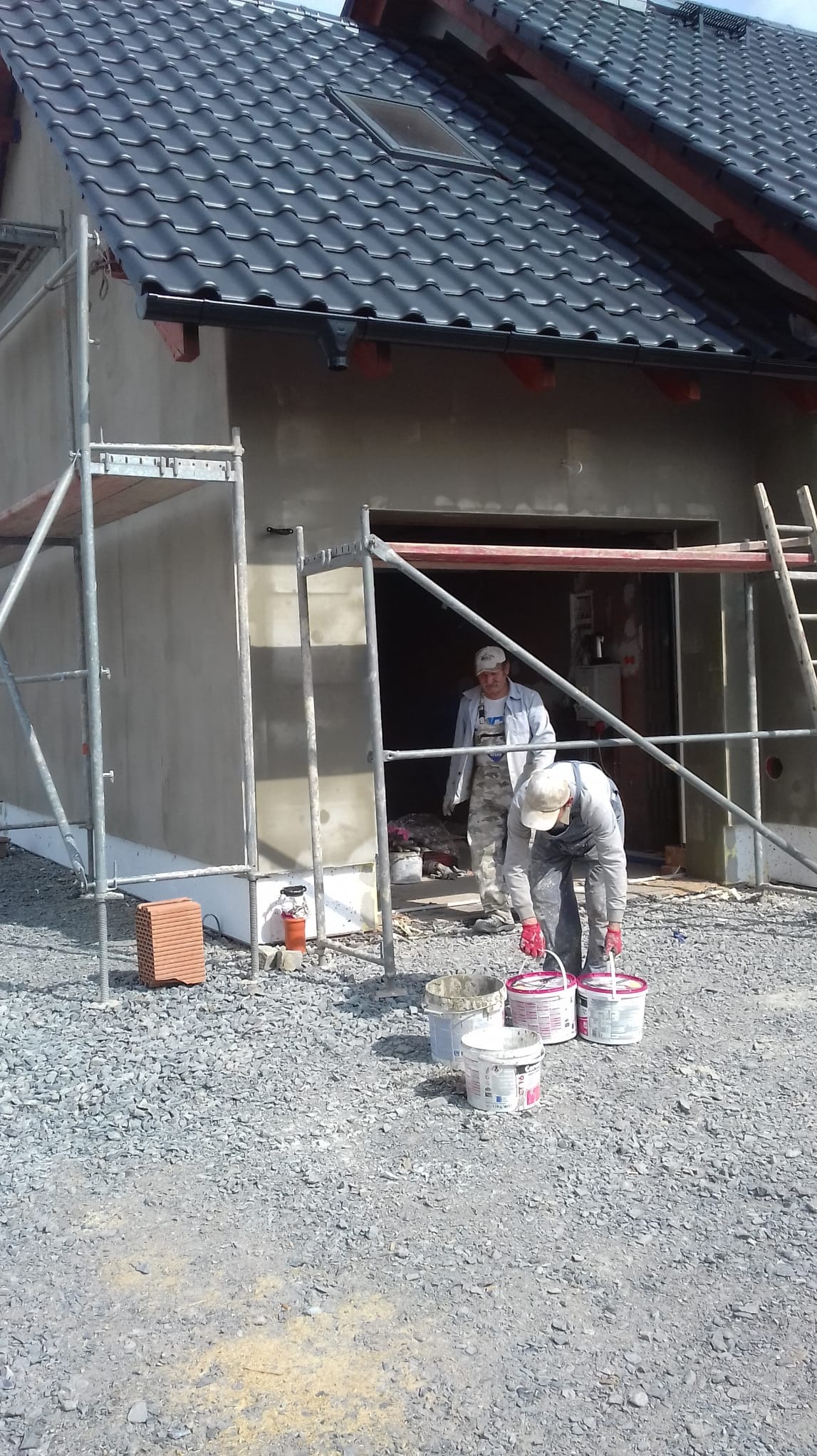 Pracownicy w trakcie przygotowań do malowania elewacji domu jednorodzinnego. Widoczne rusztowania, wiaderka z farbą i fragment otynkowanej ściany.
