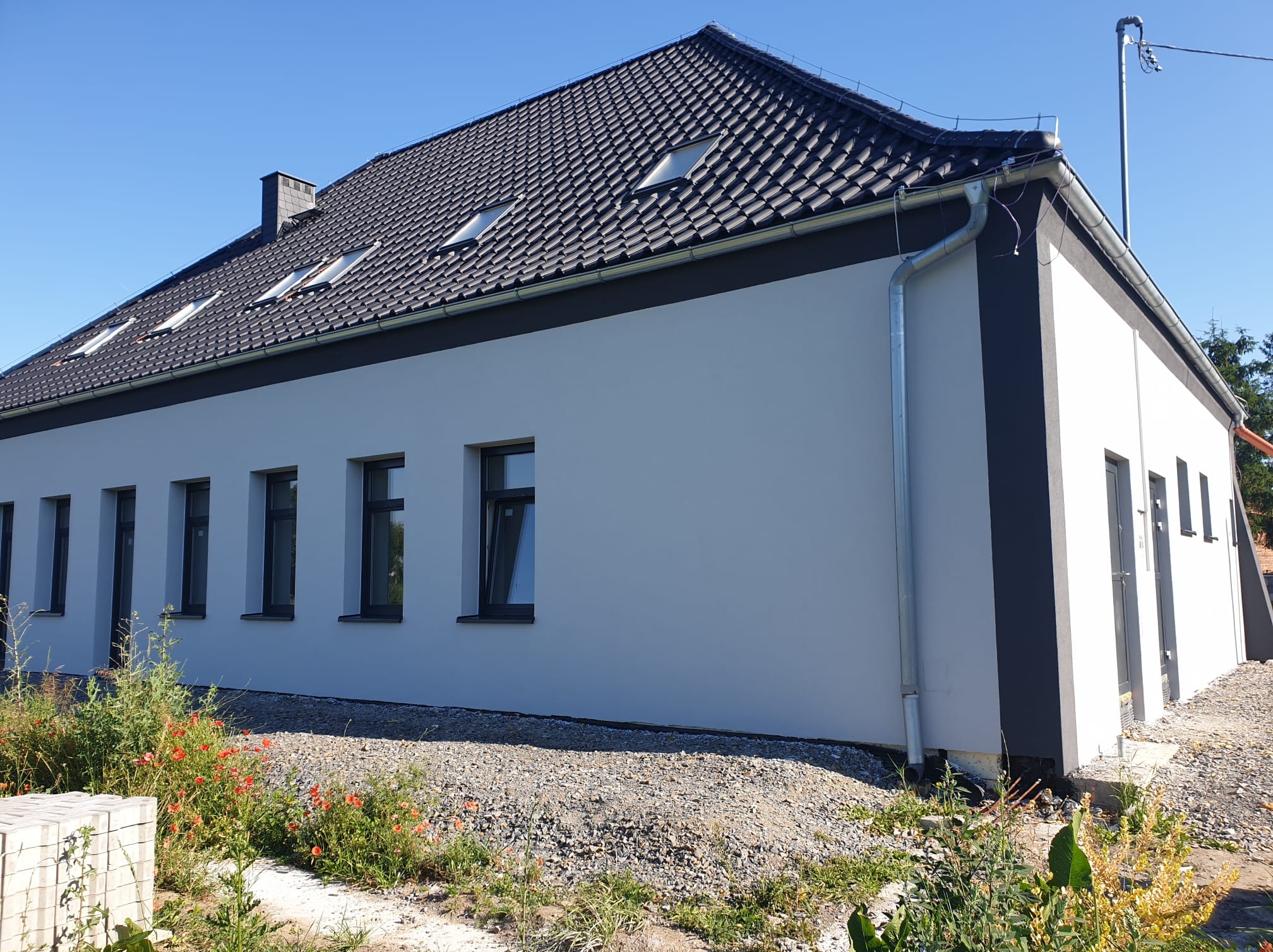 Biały budynek z ciemną dachówką i szarymi akcentami, widoczny z zewnątrz w słoneczny dzień. Widoczne okna, rynny i fragment ogrodu z roślinnością.