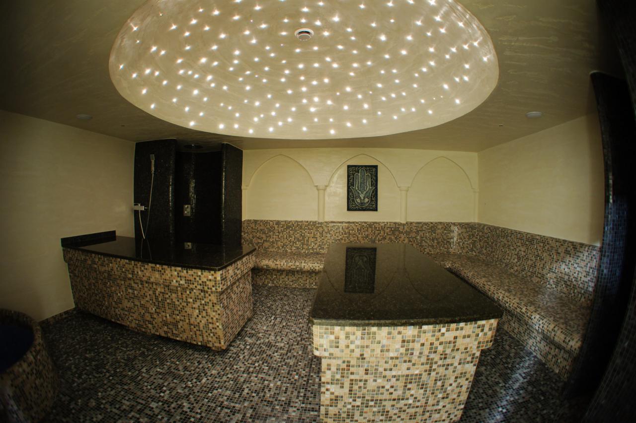 Mozaikowa sauna z oświetleniem punktowym sufitu, czarnym prysznicem i siedziskami. Dekoracyjne łuki i obraz na ścianie. Kameralna atmosfera.