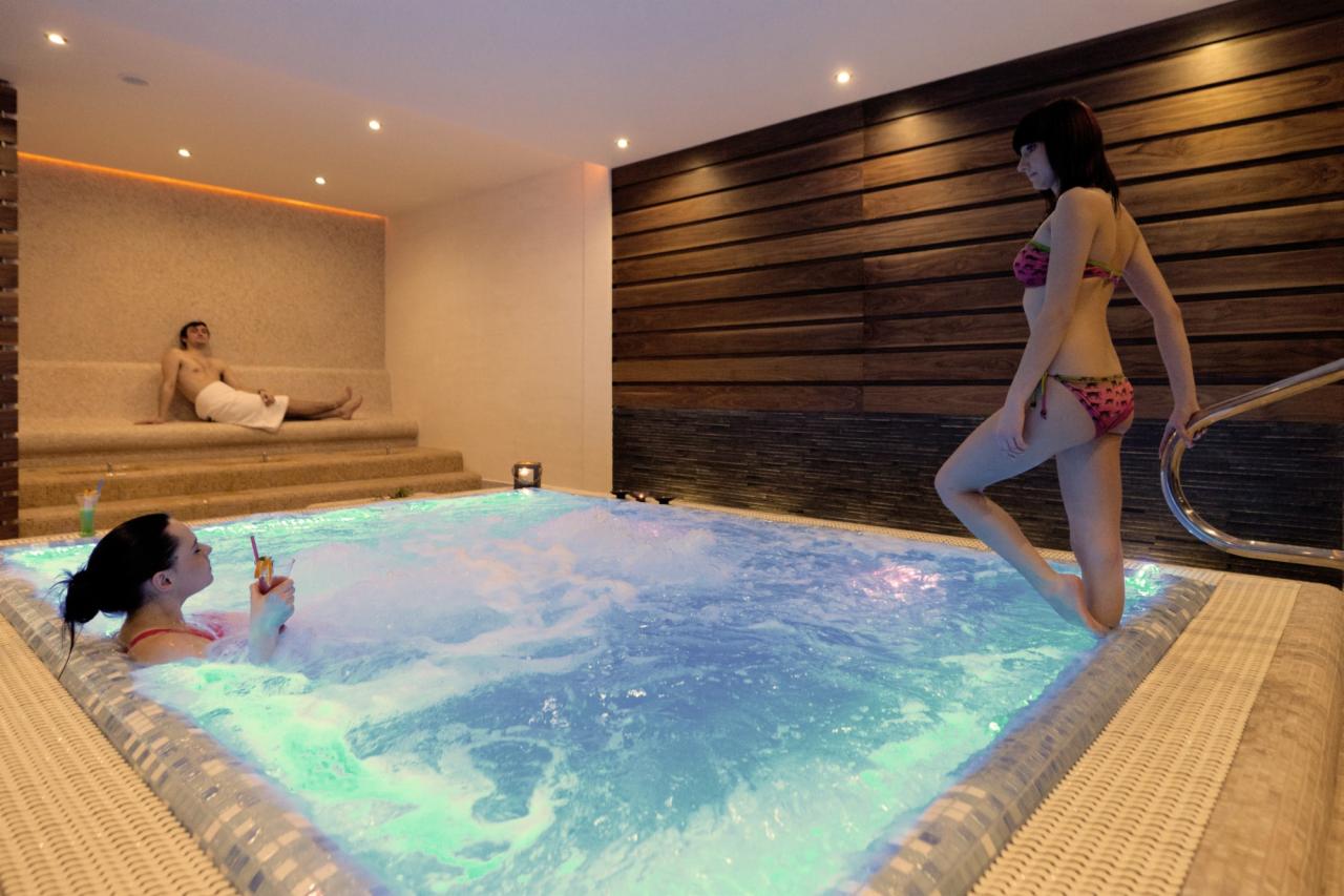 Relaks w jacuzzi: kobieta w bikini wchodzi do bąbelkowej wody, mężczyzna odpoczywa na podwyższeniu. Ciepłe, stonowane oświetlenie, drewniana ściana w tle.