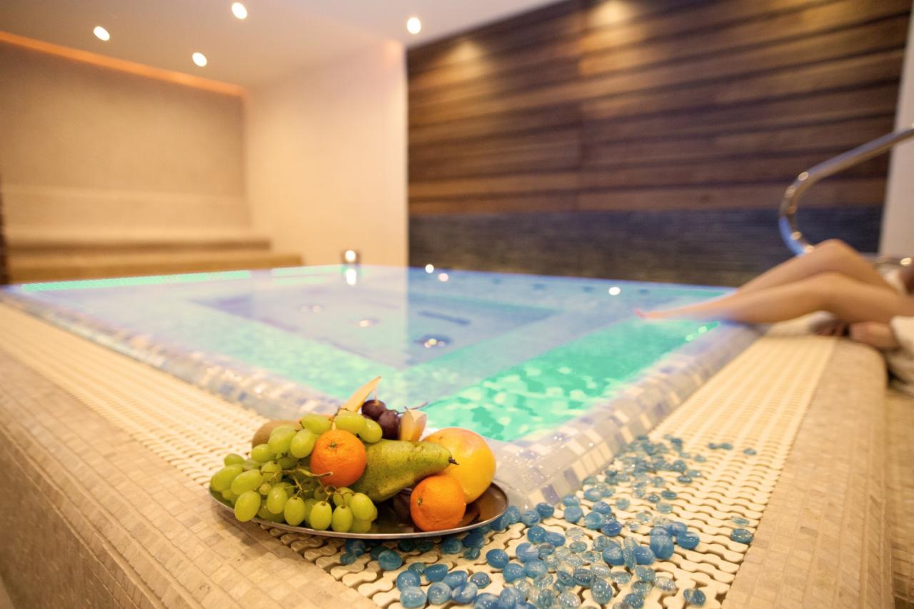 Nowoczesne jacuzzi z owocami i dekoracyjnymi kamieniami, oświetlone punktowo, z fragmentem osoby relaksującej się w tle. Drewniana ściana w tle.