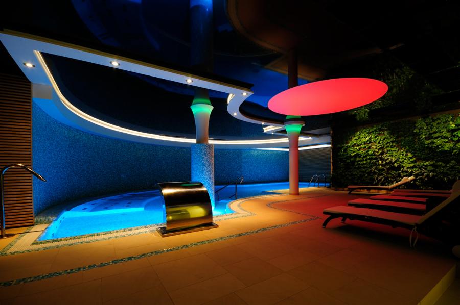 Luksusowy basen kryty z mozaiką, wodospadem i oświetleniem LED. Leżaki zachęcają do relaksu przy ścianie z zieleni. Nowoczesny design z kolumnami i podwieszanym sufitem.