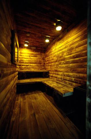 Drewniana sauna z ławkami w kształcie litery L, oświetlona punktowymi lampami sufitowymi. Widoczne deski na ścianach i podłodze, lustro na ścianie.