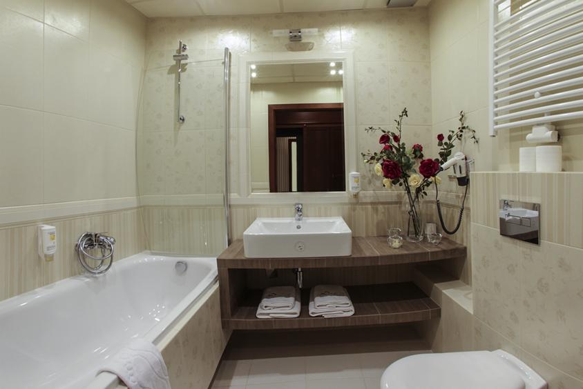 Jasna łazienka z wanną, umywalką na drewnianym blacie i dekoracyjnymi kwiatami. Widoczne płytki z delikatnym wzorem i chromowane elementy.