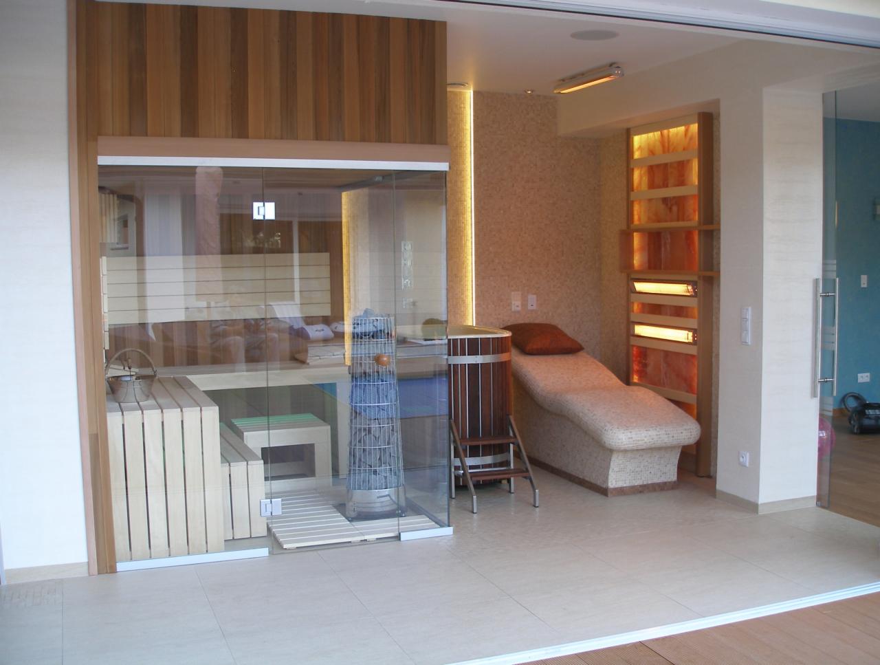 Nowoczesna sauna z przeszklonymi ścianami, drewnianymi ławkami i piecem. Obok leżanka i dekoracyjna ściana z podświetlonymi panelami solnymi. Jasne płytki podłogowe.