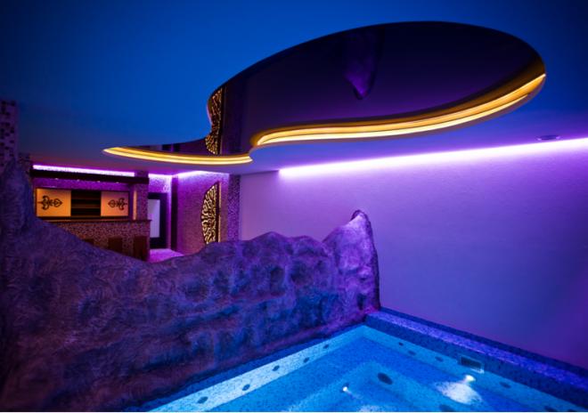 Nowoczesny basen wewnętrzny z dekoracyjnym oświetleniem LED i elementami imitującymi skałę, podświetlony na fioletowo. Widoczny bar w tle.