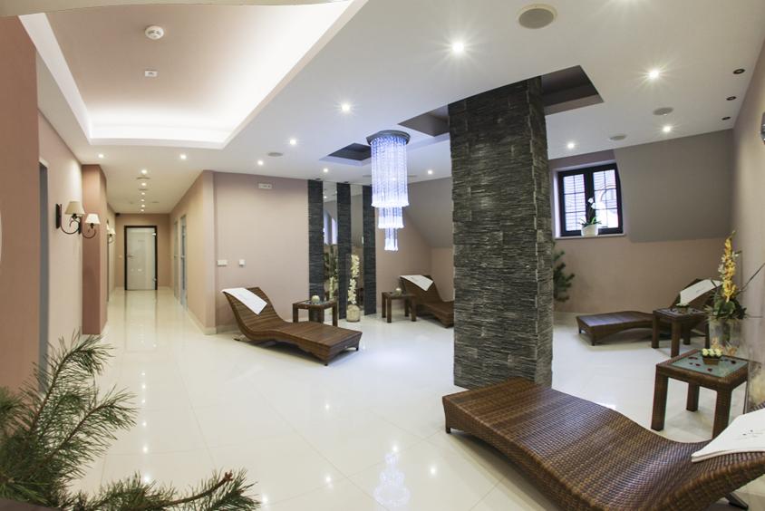 Wnętrze luksusowej sauny z leżankami z rattanu, kolumną z kamienia i kryształowym żyrandolem. Jasne płytki podłogowe i subtelne oświetlenie tworzą relaksującą atmosferę.