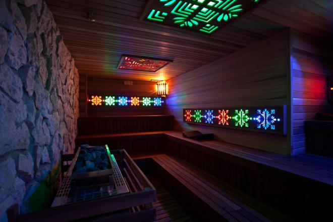Wnętrze sauny z drewnianymi ławami, oświetleniem LED i ścianą z kamienia. Widoczny piec z kamieniami i dekoracyjne panele świetlne.