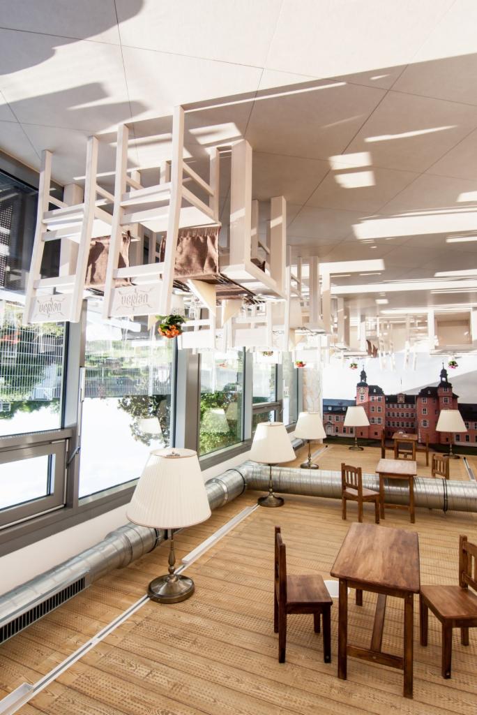 Nietypowe wnętrze kawiarni z odwróconymi meblami pod sufitem, lampami stołowymi i widokiem na zewnątrz. Uwagę przyciąga dekoracja ściany z motywem zamku.