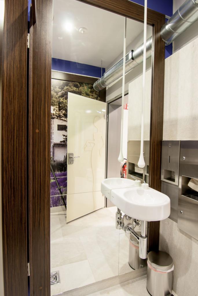 Nowoczesna łazienka z podwójną umywalką na tle drzwi z grafiką kobiety i widokiem na zewnątrz. Widoczna wentylacja i metalowe akcesoria.