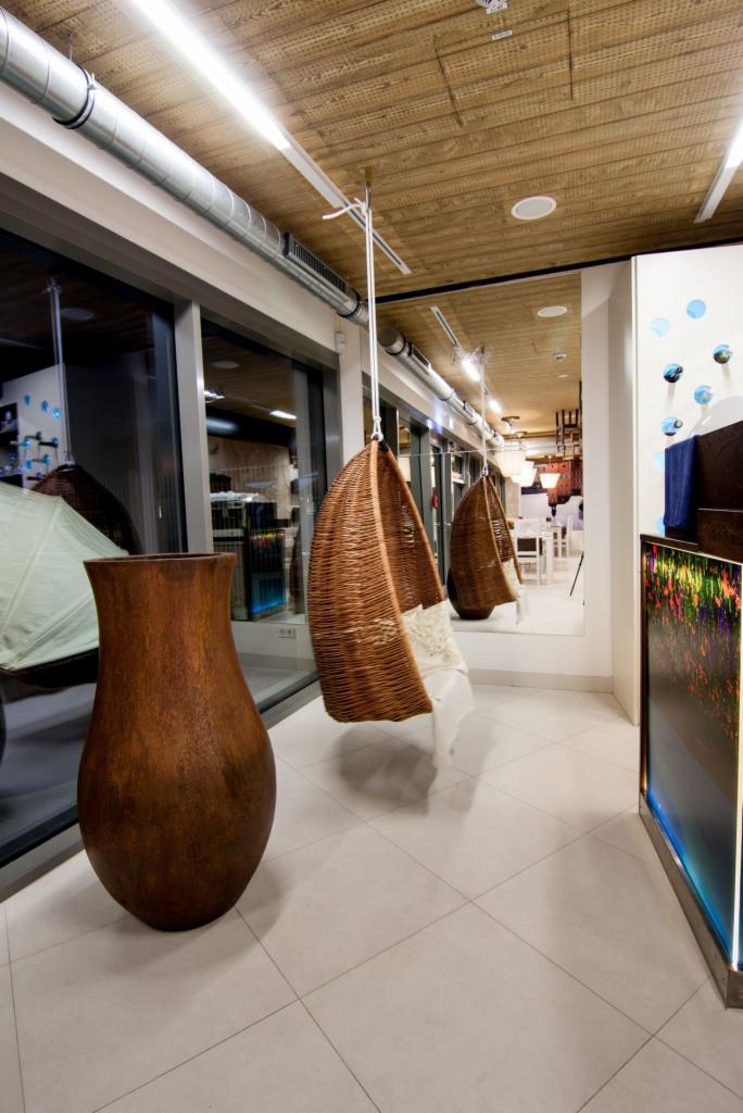 Nowoczesne wnętrze z wiklinowymi huśtawkami, dużą glinianą wazą i lustrem. Widoczne elementy instalacji pod sufitem i płytki podłogowe ułożone w karo.