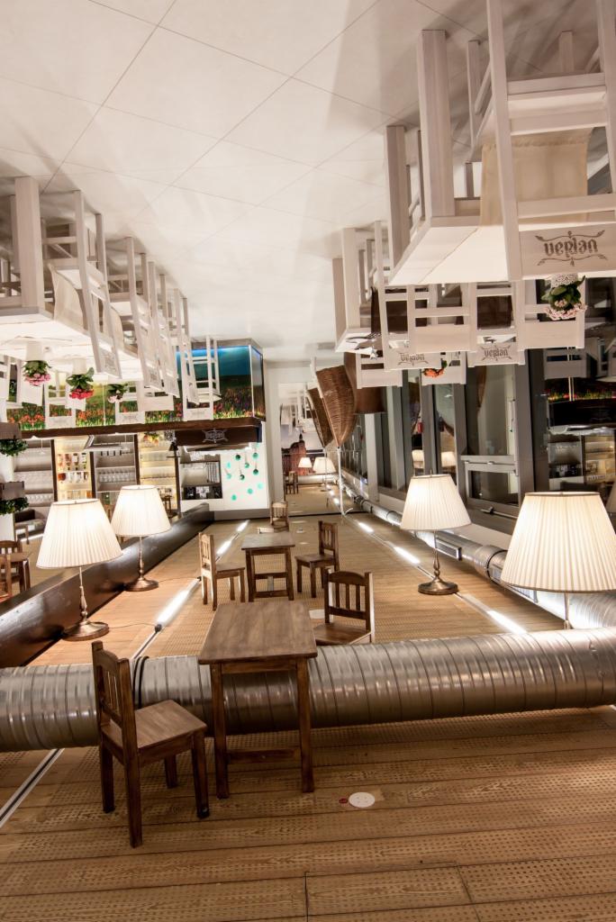 Awangardowa aranżacja wnętrza restauracji z odwróconymi krzesłami pod sufitem i nietypowym oświetleniem. Widoczne stoliki i akwarium z roślinami.