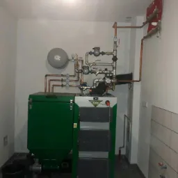 Kotłownia z zielonym piecem na paliwo stałe, instalacją z miedzianych rur, zbiornikiem wyrównawczym i rozdzielaczem na ścianie w pomieszczeniu wyłożonym częściowo białymi płytkami.