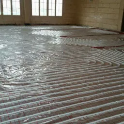 Białe rurki układane spiralnie na czerwonej siatce pod folią, przygotowanie do wylewki pod system grzewczy w jasnym pomieszczeniu z oknami.