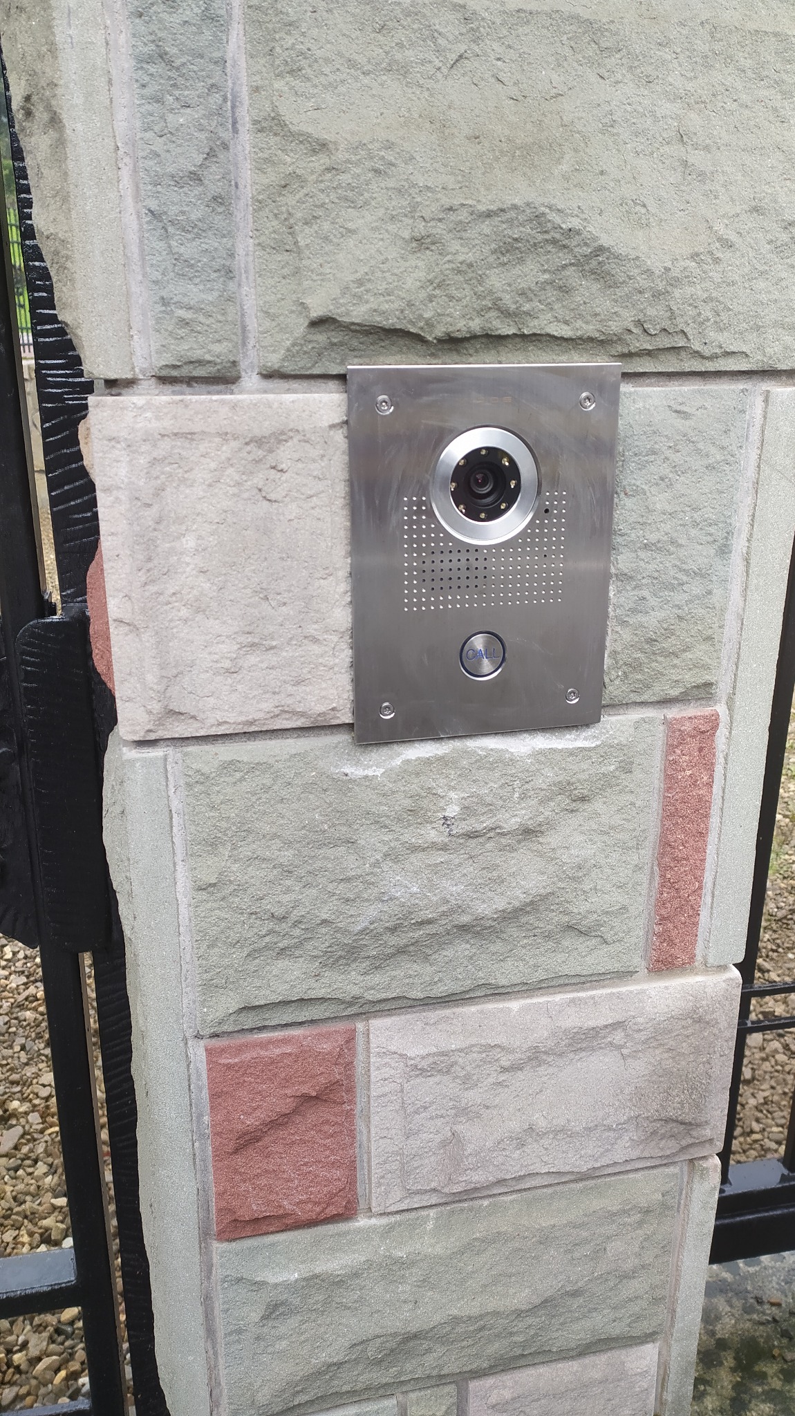 Stalowy wideodomofon z kamerą na murowanym słupku ogrodzenia z kamienia naturalnego. Widoczny przycisk dzwonka i fragment czarnego, metalowego ogrodzenia.