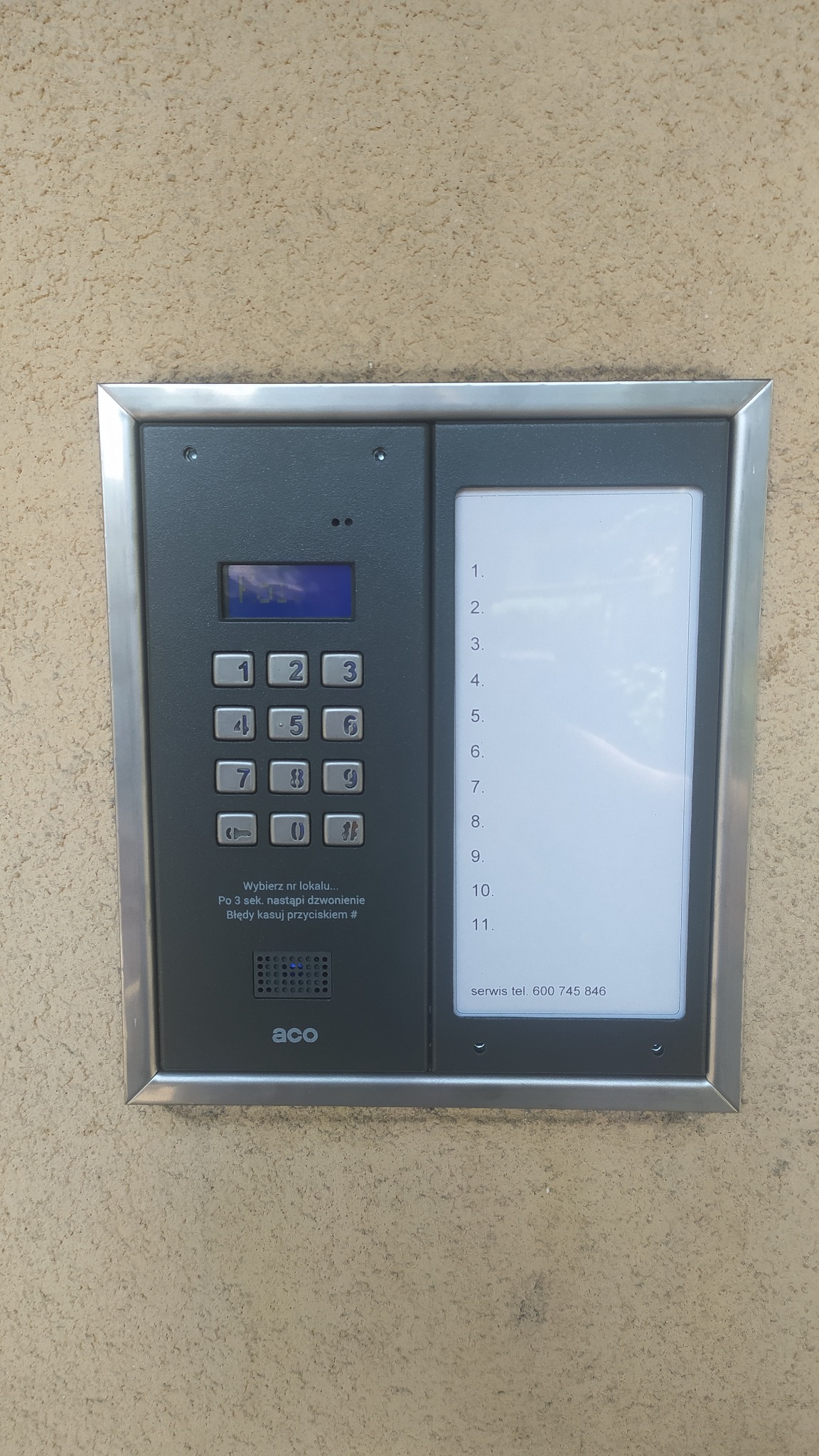 Zamontowany panel domofonowy ACO z klawiaturą numeryczną i listą lokatorów na elewacji budynku. Widoczne przyciski i wyświetlacz.